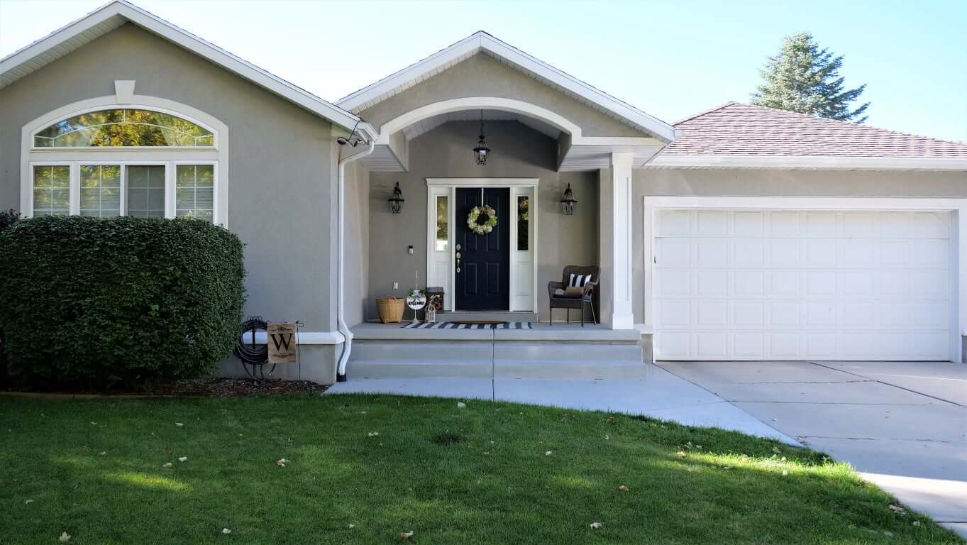 Stucco home with a covered front porch and concrete steps down to the driveway, two-car garage, and green lawn.