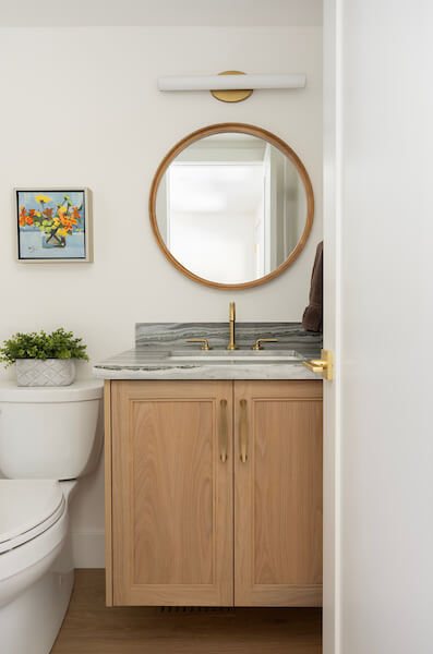 Simple powder bathroom design with a vanity and toilet next to each other on the wall. Above the vanity is a round mirror and modern, horizontal light fixture.