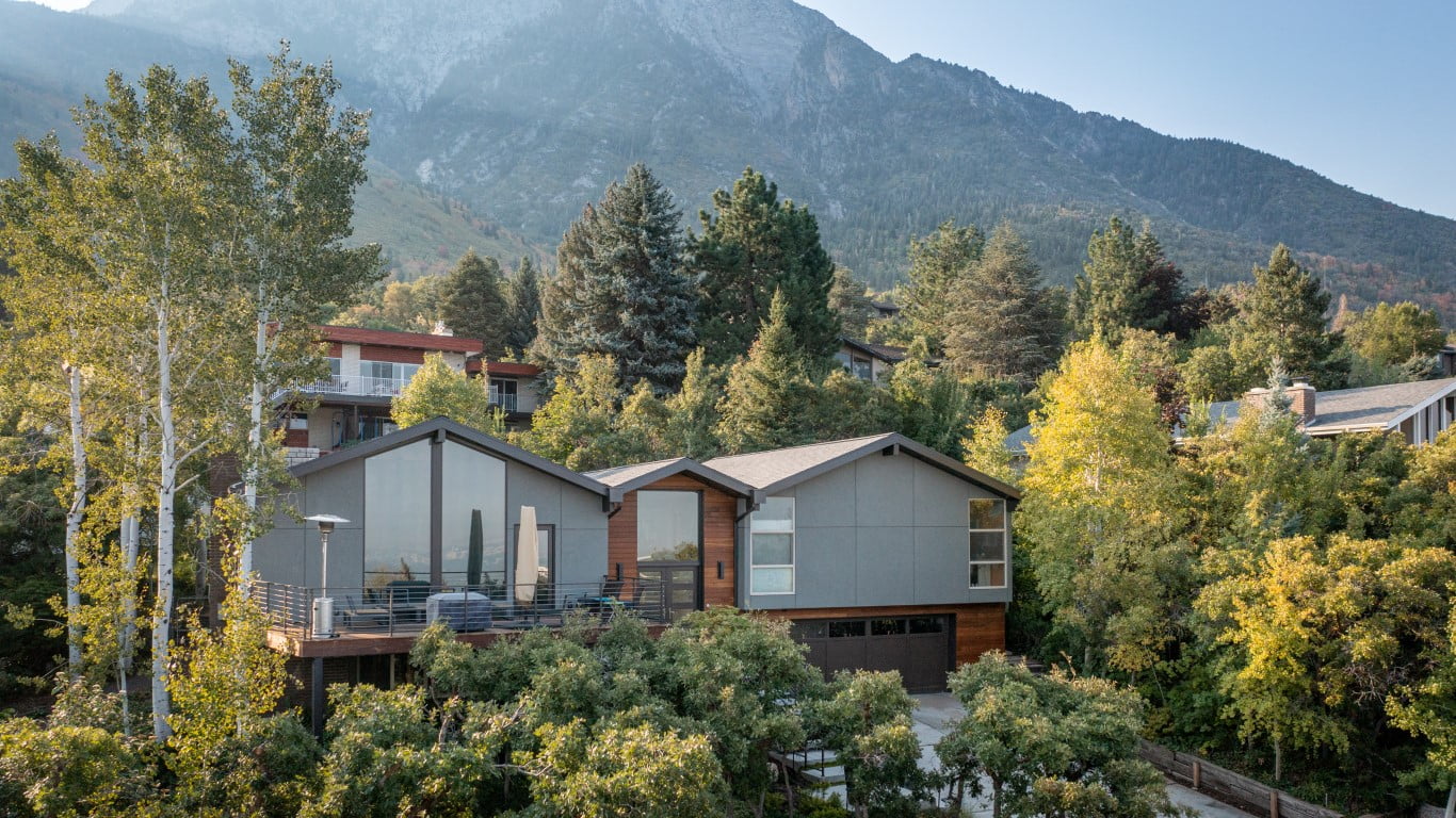 Exterior of split level home after the renovation with a two-car garage, updated landscaping, and a modern railing leading up to the front porch and on the deck. The home sits in front of a beautiful mountain backdrop and is partially hidden by the landscaping.