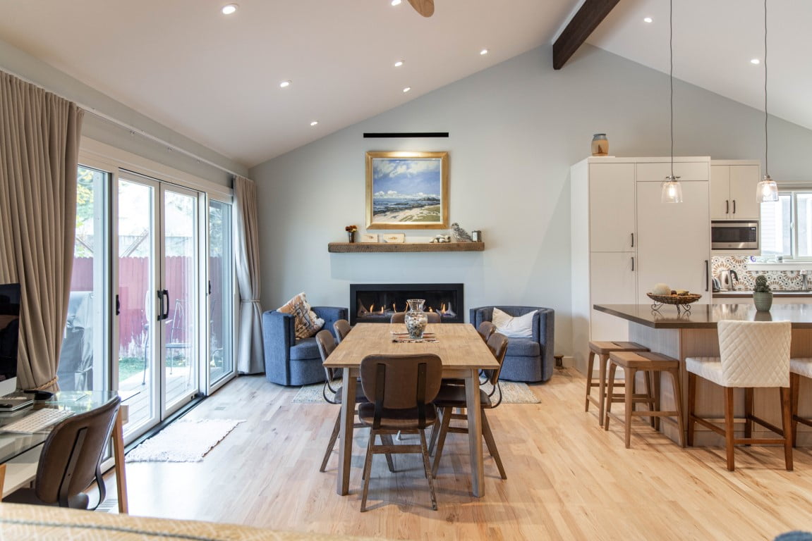 Renovated open concept great room with vaulted ceilings. The dining table sits in front of the modern fireplace and has a table with seating for six. On the left are glass sliding doors to the patio. On the right is a small view to the updated kitchen.