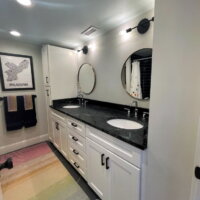Thumbnail of http://Bathroom%20with%20double%20sinks%20and%20a%20storage%20cabinet%20on%20the%20end.%20Above%20both%20sinks%20are%20round%20mirrors%20and%20light%20fixtures.%20In%20front%20of%20the%20vanity,%20on%20the%20floor,%20is%20a%20colorful%20rug.