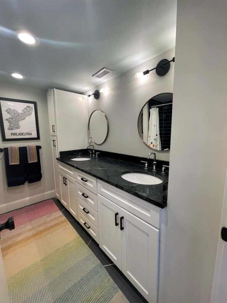Bathroom with double sinks and a storage cabinet on the end. Above both sinks are round mirrors and light fixtures. In front of the vanity, on the floor, is a colorful rug.