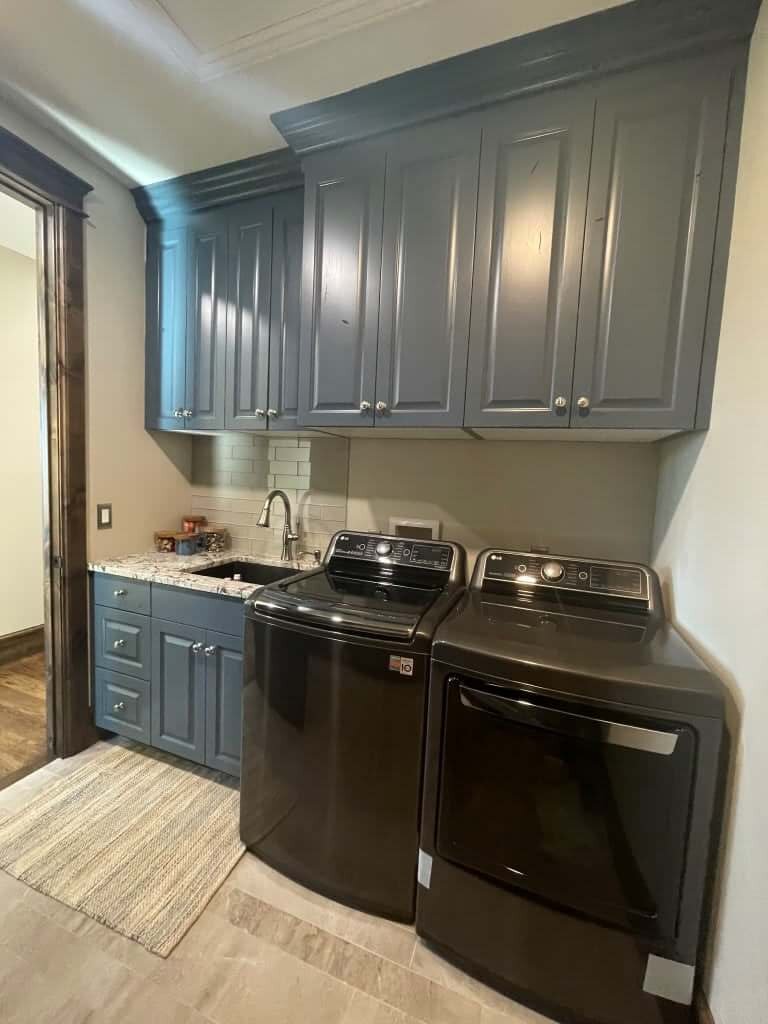 Laundry room with side-by-side washer and dryer, blue painted cabinets, and a sink with a small area rug on the floor in front.