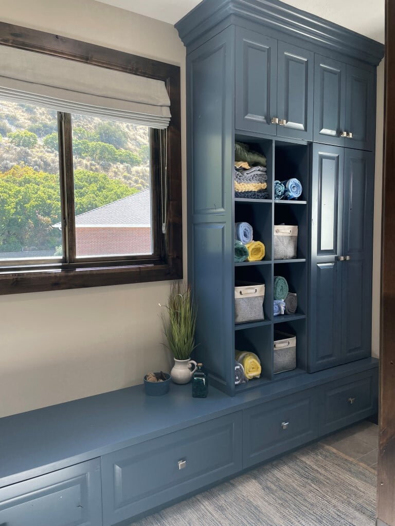 Built-in cabinets painted blue. The built-ins include open shelving with baskets and rolled towels, a bench seat under the windows, and cabinets.