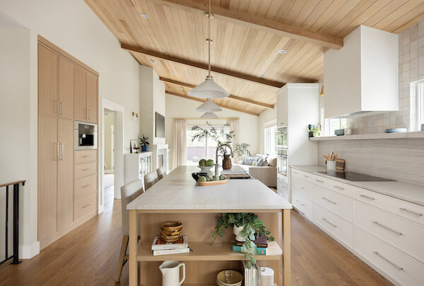 Open concept kitchen with a large island in the center. On the right is a wall of cabinetry that includes the oven, stove, and fridge. In the center is a large island with open shelves on the end , the sink, and seating. On the left is a additional cabinetry with a built-in coffee machine.