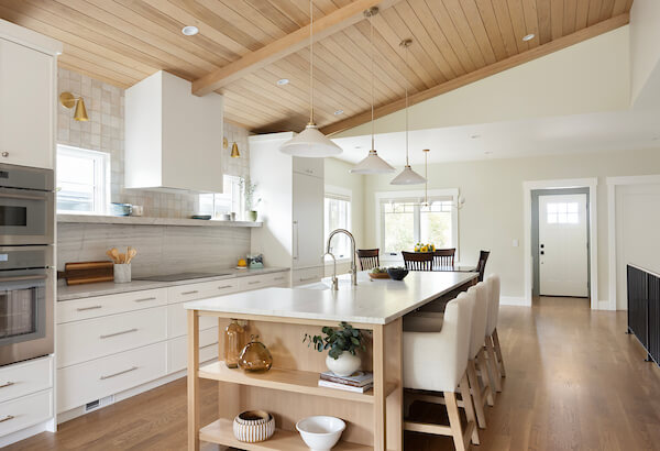 Open concept kitchen with a large island in the center. On the left is a wall of cabinetry that includes the oven, stove, and fridge. In the center is a large island with open shelves on the end , the sink, and seating.