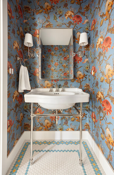 Powder bathroom vanity. The console style sink is centered on the wall with a mirror and sconces above. On the left is a towel hook. The spaces features an elegant floral wallpaper and small hexagon tiles on the floor.
