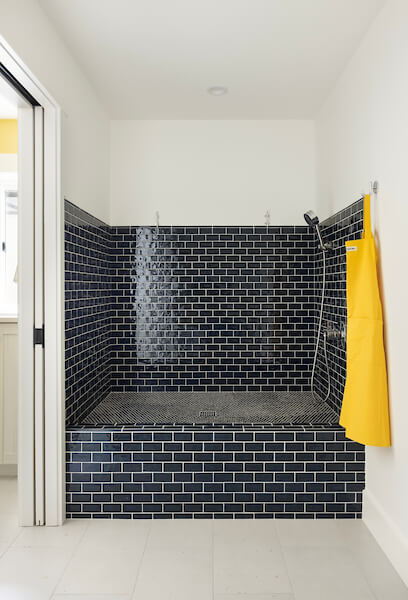 At-home dog wash station. The dog wash is elevated from the floor and features blue tile. On the wall to the left is a pocket door, straight ahead is the dog wash, and on the right is a hook with a yellow apron.