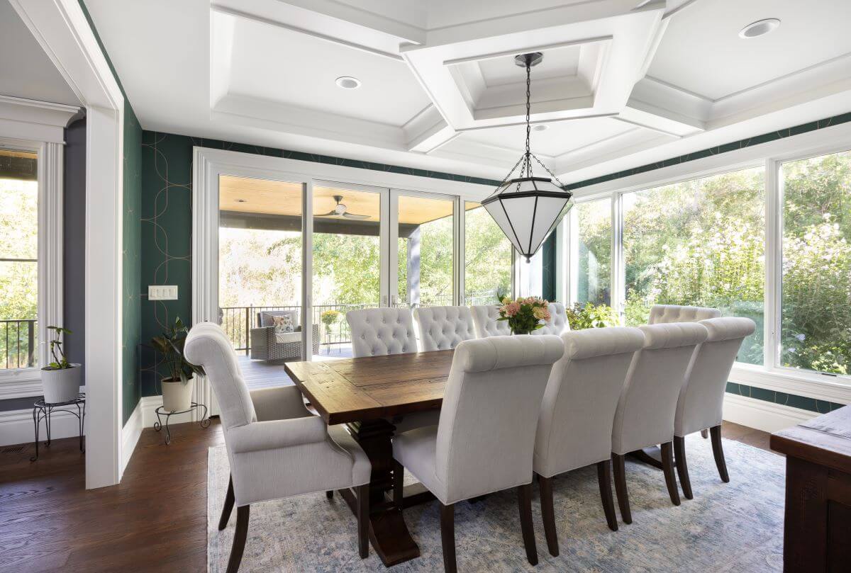 Dining room with large windows, doors to the covered deck, a green wallpaper design on the walls, and a table for 10 in the center.