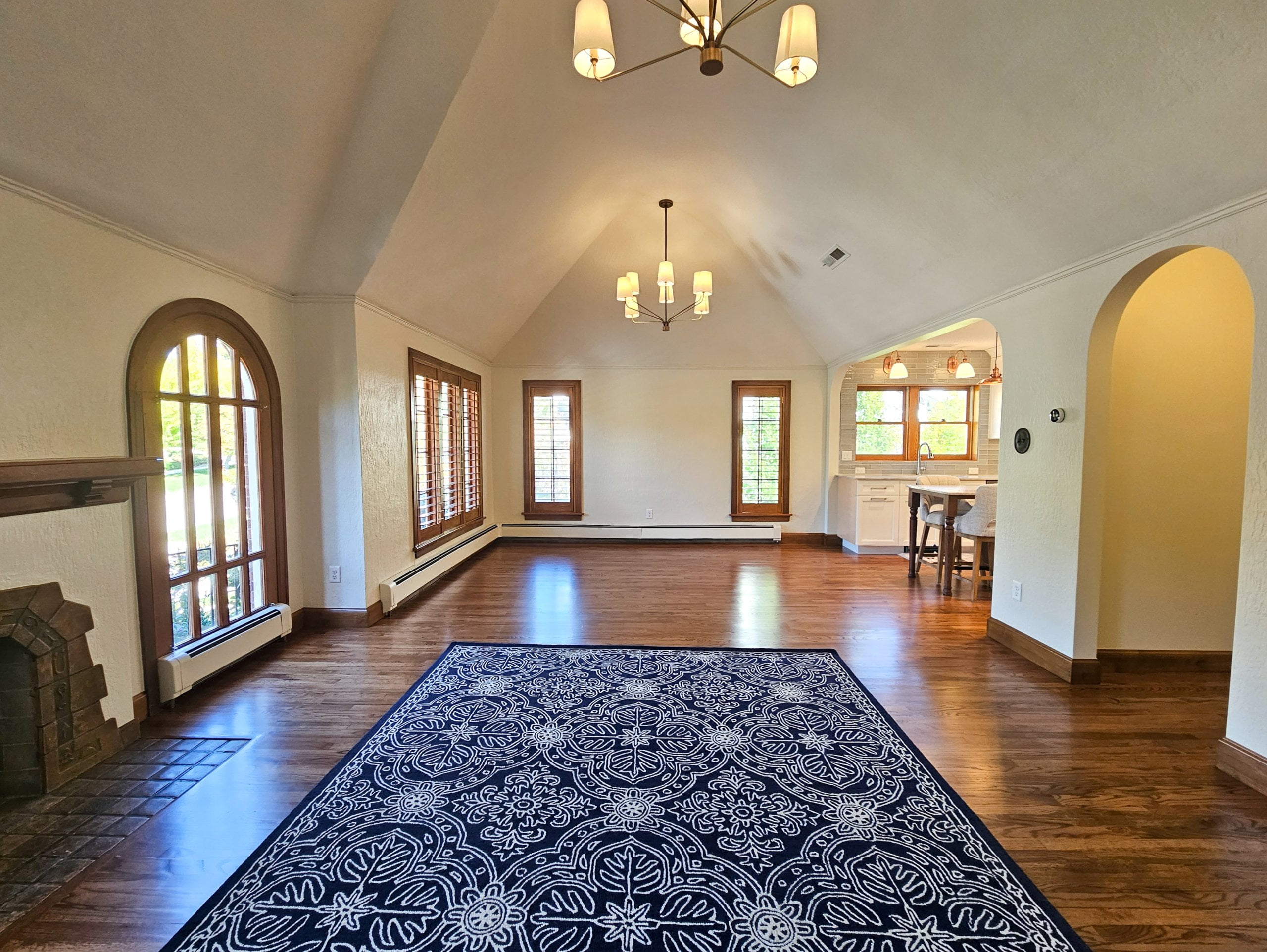 Historic home great room remodel with vaulted ceilings, an open concept living room and dining room, and arch openings with access to the hallway and kitchen. In the living room a large area rug is placed on the floor. In the living room and dining room identical lights are hung from the vaulted ceiling.