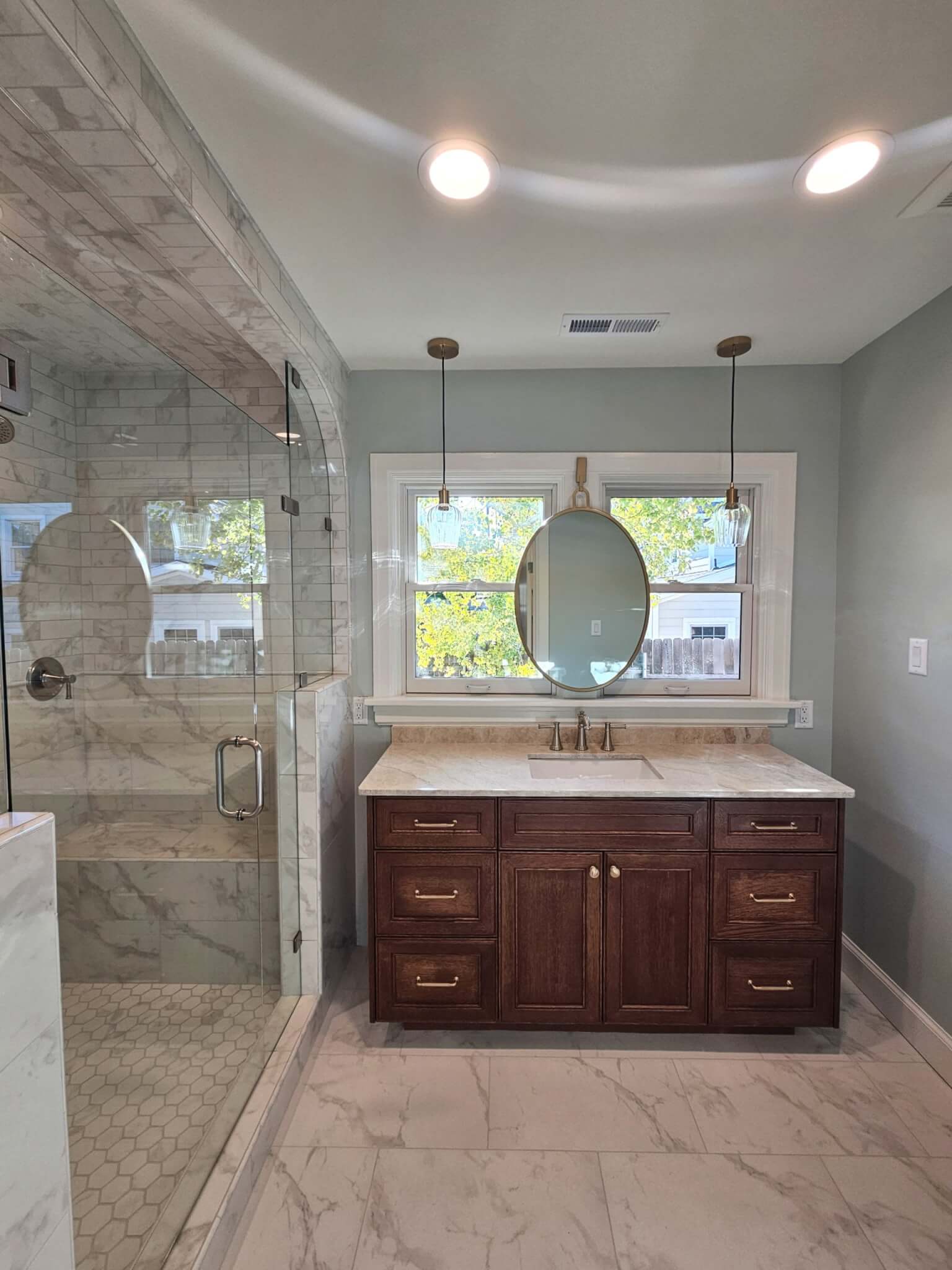 Bathroom sink and shower. The sink is located in front of the two windows with a mirror centered between and lights hung from the ceiling on either side. The shower is enclosed with glass and features a bench.